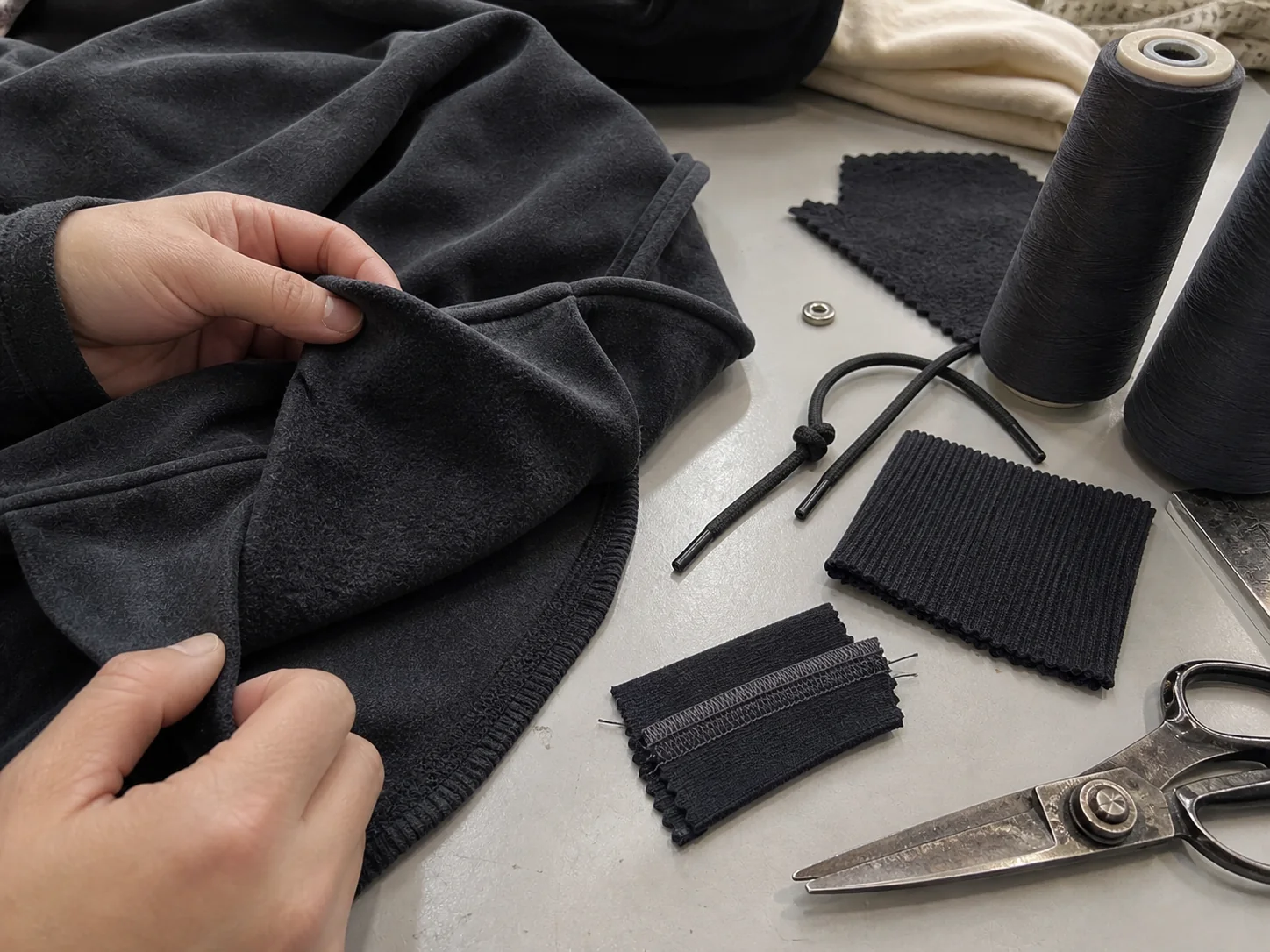 Hands comparing charcoal fleece, rib knit, drawcord and seam construction samples