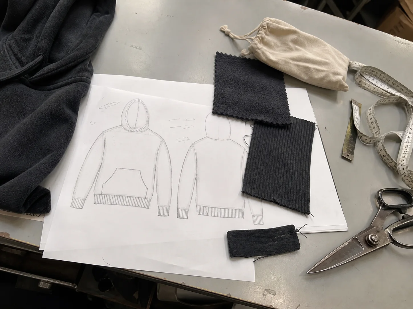 Charcoal hoodie technical sketch with fleece and rib fabric swatches on a sample-room table